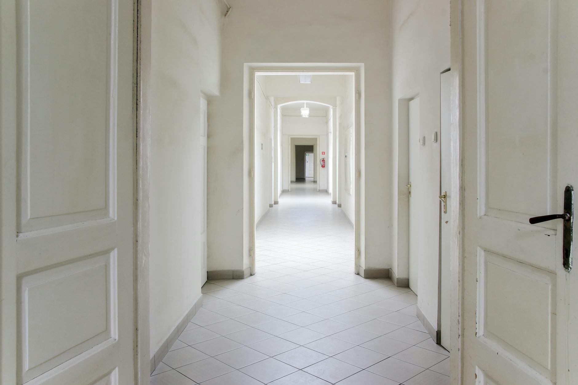 A long, white empty corridor with open doors — the threshold of irreversible decisions.